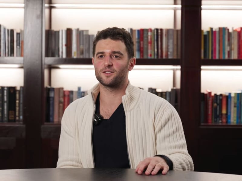 Austin Rief seated at a table in a cream ribbed jacket, speaking in front of a lit bookshelf